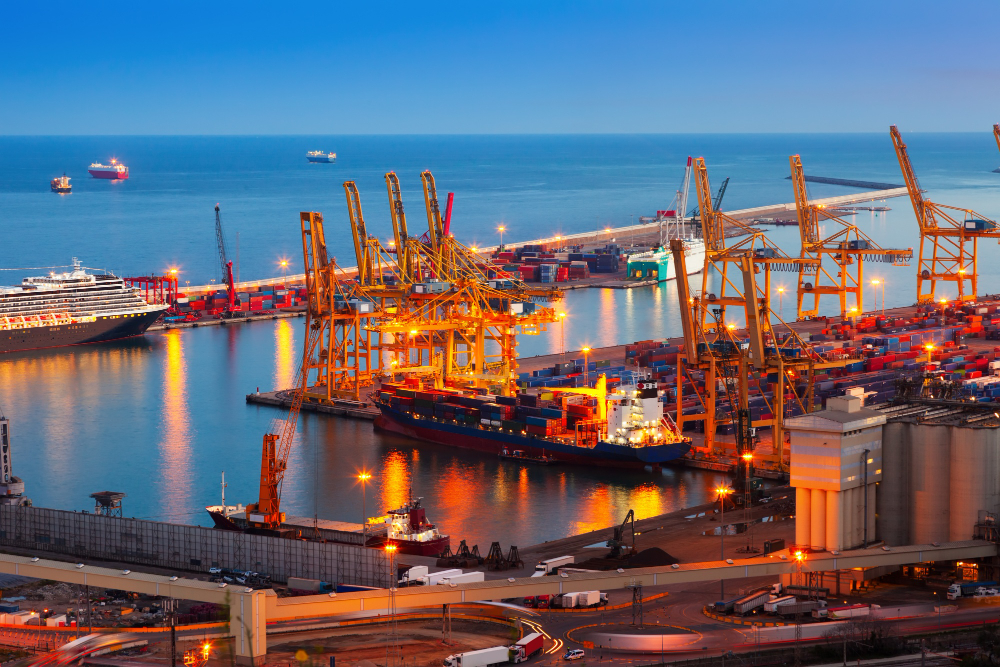 Busy shipping port at dusk with yellow gantry cranes, containers, and ships docked along a calm sea.