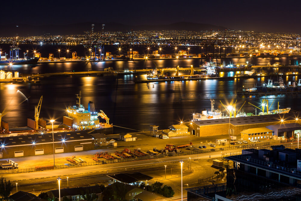 Nighttime port scene with lit cranes, cargo ships, and a city skyline in the distance.